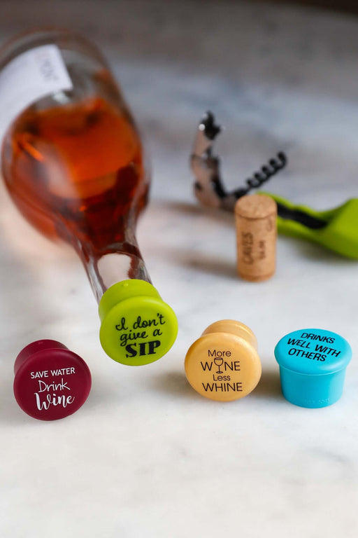 Silicone wine caps with humorous messages on a marble surface, alongside a bottle of wine and corkscrew.