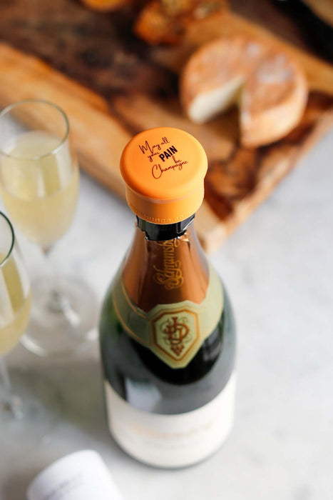 Champagne bottle with orange stopper inscribed 'May All Your Pain Be Champagne' on a wooden cutting board.