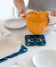 Person holding an orange pot over an interlocking silicone trivet on a dining table with plates.