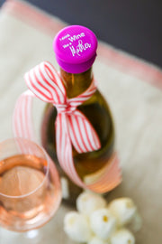I Drink Wine Like a Mother silicone cap on a wine bottle with a ribbon, beside a glass of rosé wine.