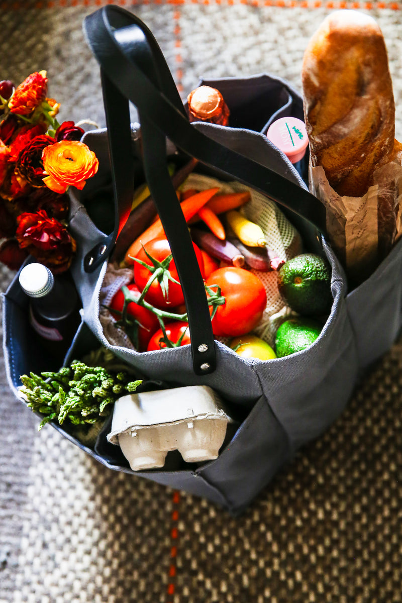 A grey multi-pocketed market tote filled with fresh vegetables, eggs, and a loaf of bread, ideal for grocery shopping.