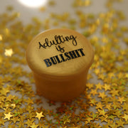 Adulting is Bullshit silicone wine cap on a background of gold star confetti.