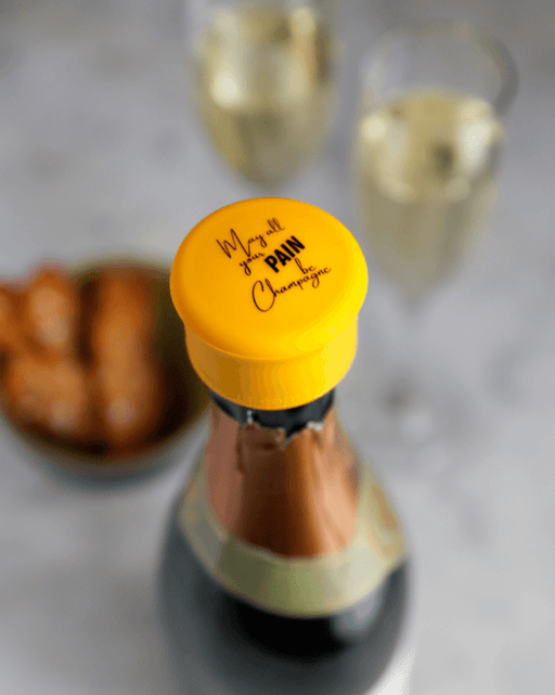 May All Your Pain Be Champagne bottle with reseal champagne stopper and glasses of sparkling wine.