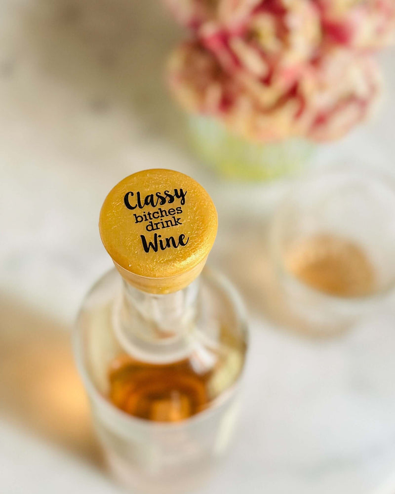 Top view of Classy Bitches Drink Wine silicone cap on a wine bottle with blurred background of flowers.