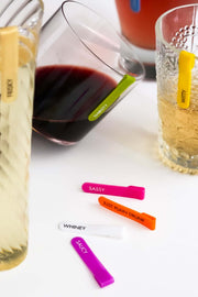 Colorful mini silicone identifiers for stemless glasses, featuring labels like 'Sassy' and 'Whiney', on a table.