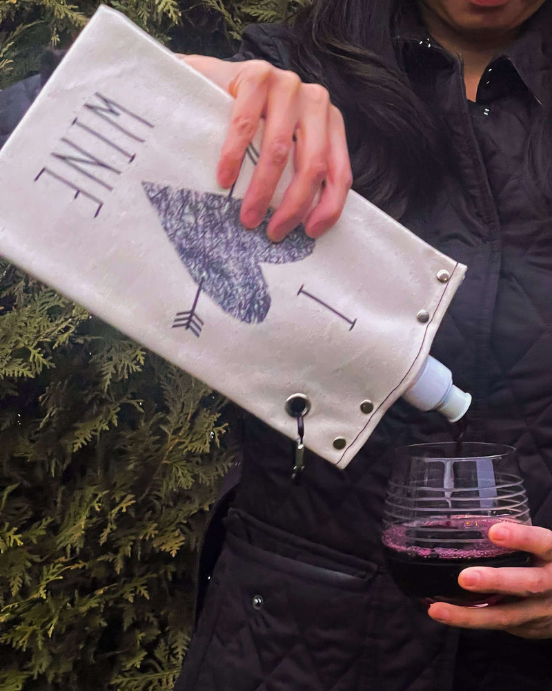 Person pouring wine from I Heart Wine canvas canteen into a glass, showcasing its stylish design and functionality.