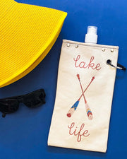 Paddle Lake canvas canteen with 'lake life' design, sunglasses, and yellow sun hat on a blue background.