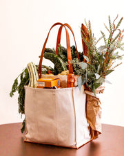 Natural multi-pocket canvas market tote filled with groceries and greenery on a wooden table.