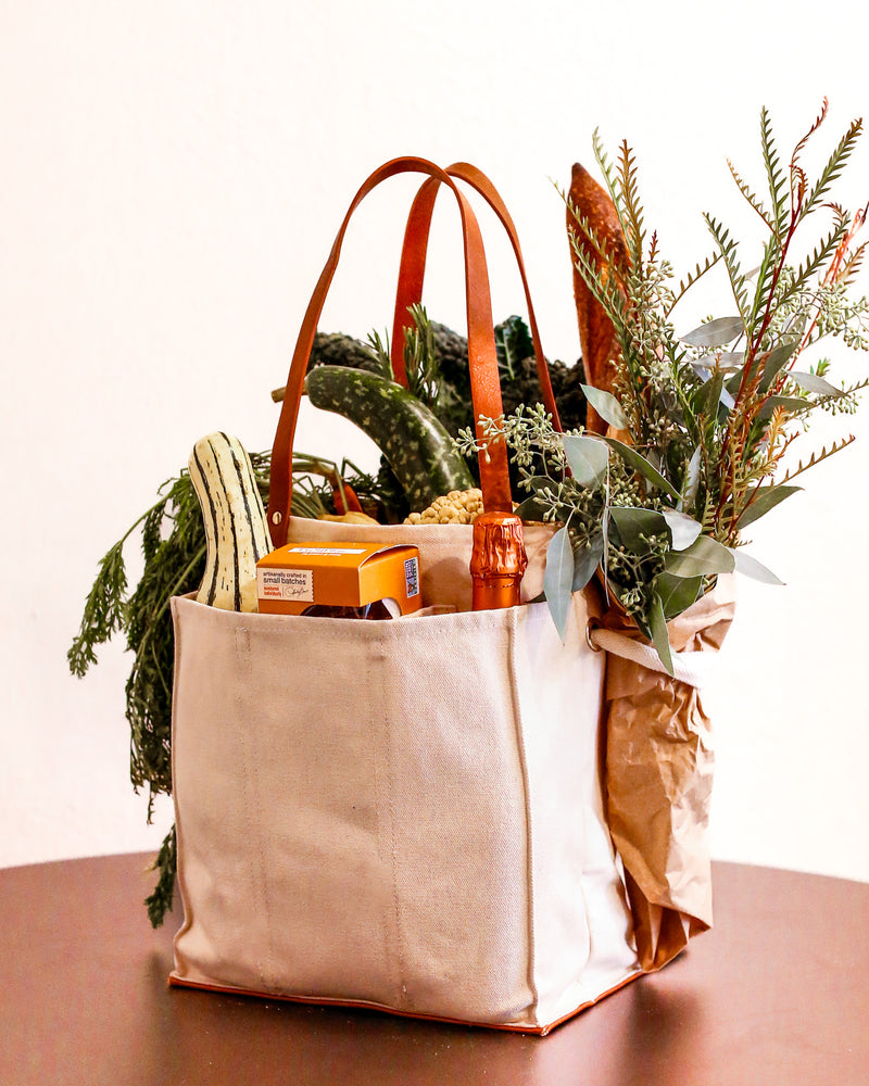 Natural multi-pocket canvas market tote filled with groceries and greenery on a wooden table.