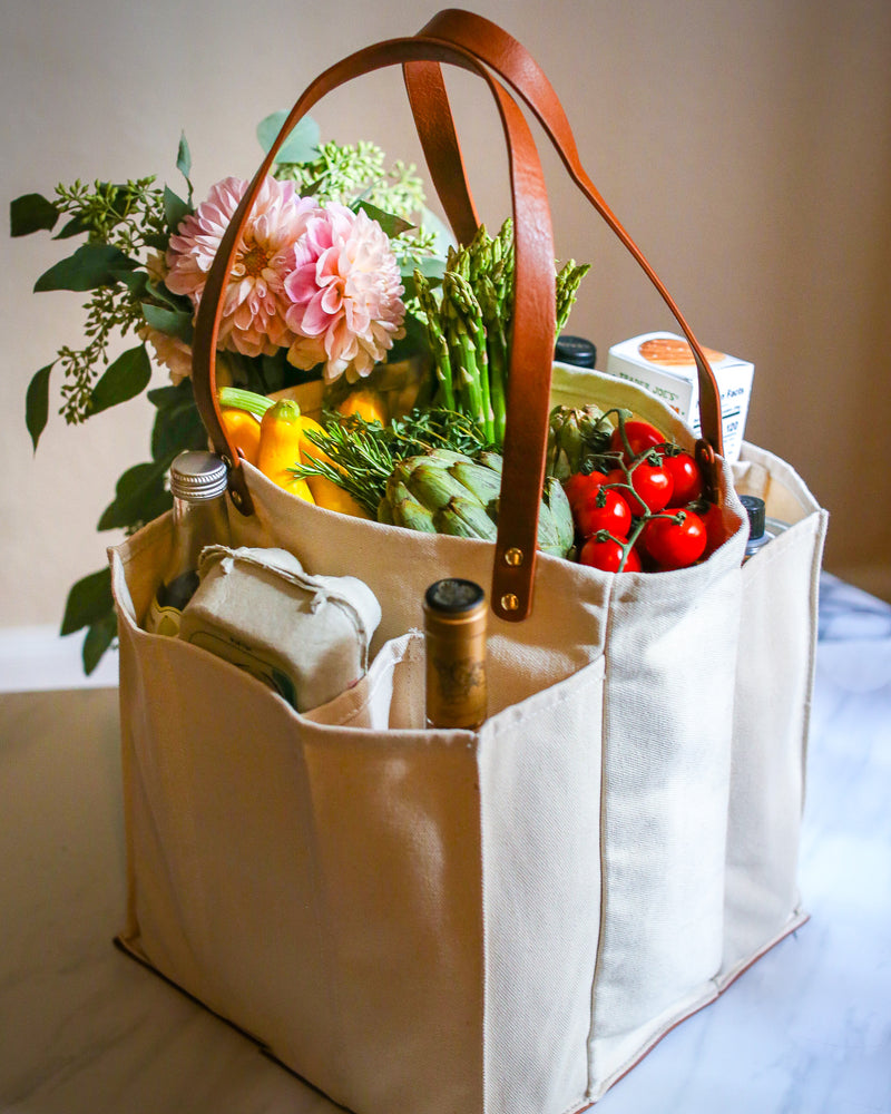 Natural multi-pocket canvas market tote filled with fresh groceries and flowers, featuring sturdy leather handles.