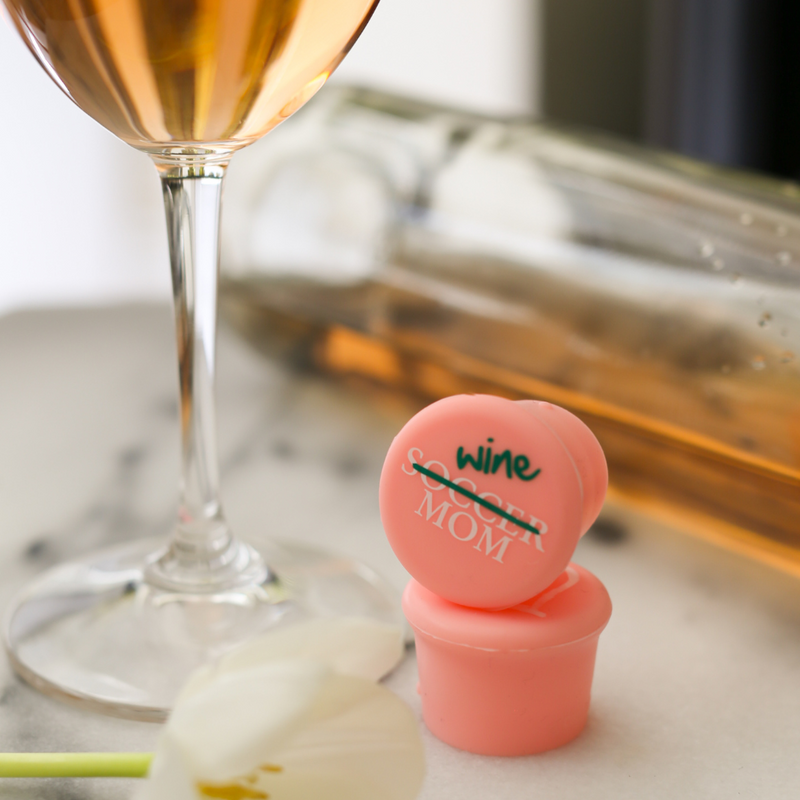 Silicone wine cap designed for soccer moms next to a glass of wine, perfect for resealing bottles.