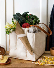 Natural multi-pocket canvas market tote filled with fresh vegetables, flowers, and groceries on a wooden table.