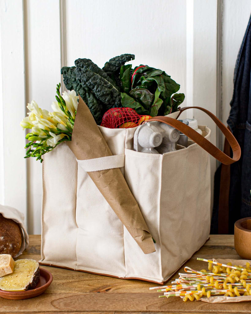 Natural multi-pocket canvas market tote filled with fresh vegetables, flowers, and groceries on a wooden table.