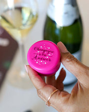 Pink Champagne Stopper with 'Champagne is the Answer' text held in hand, with wine glass and bottle in background.