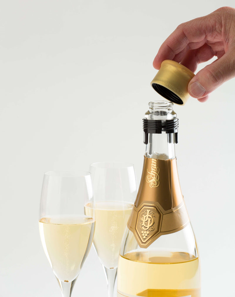Hand placing a golden champagne stopper on a bottle with two glasses of sparkling wine.