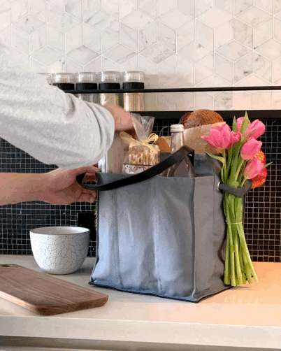 Person using a grey multi-pocketed market tote to organize groceries and flowers on a kitchen counter.