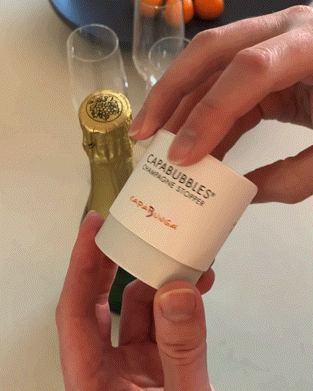 Hand holding CapaBubbles Champagne Stopper beside a champagne bottle, perfect for preserving sparkling wine.