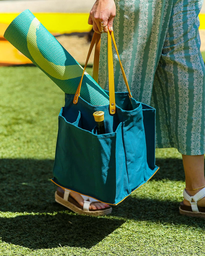 Person carrying a petrol blue market tote with multiple pockets, holding a yoga mat and a bottle, on green grass.